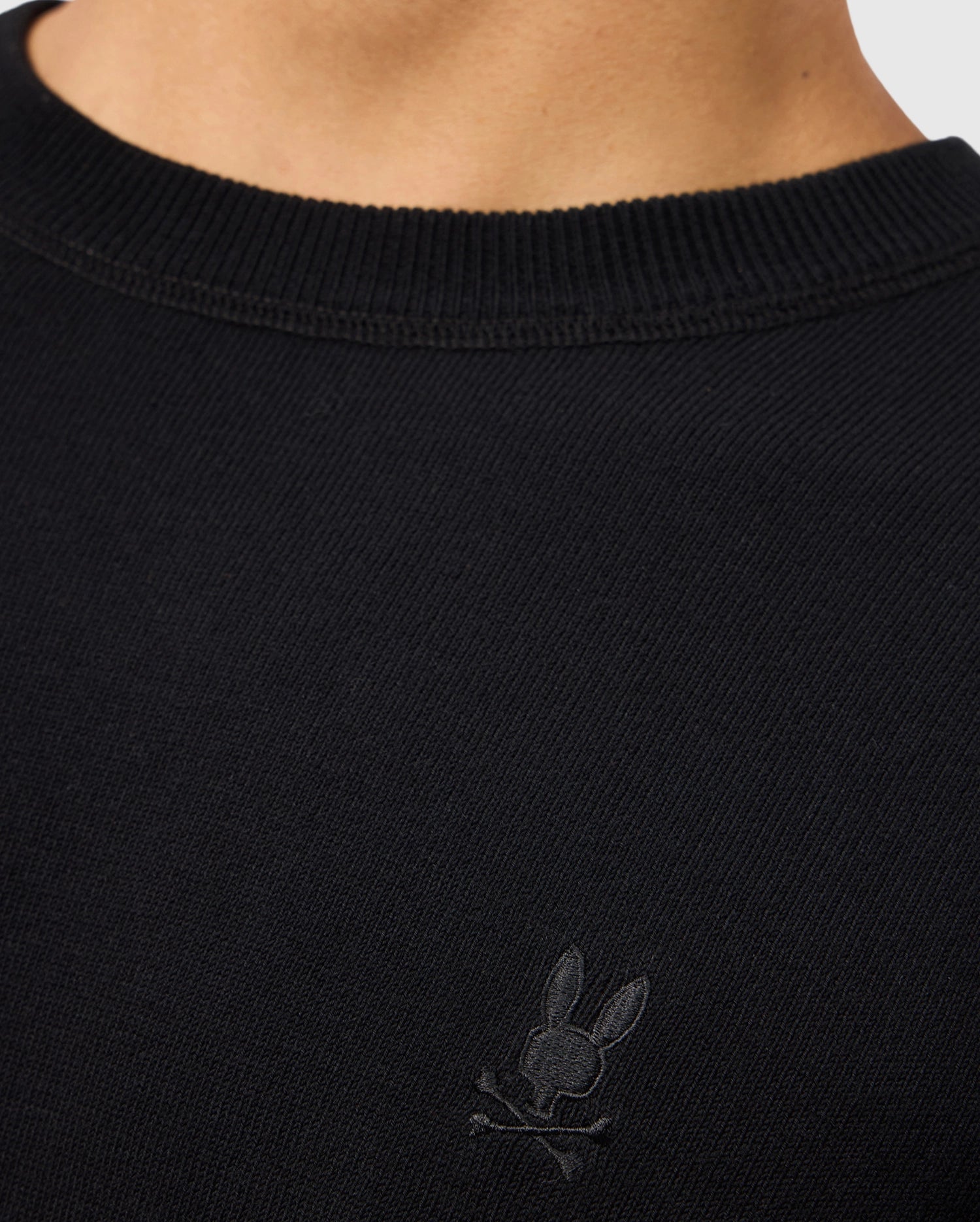 The image showcases a person wearing the Psycho Bunny MENS BELMORE EXTRA FINE COTTON SWEATER - B6E832D200, which features an embroidered bunny skull and crossbones logo on the chest. Made from extrafine cotton, this black sweater includes a ribbed neckline that highlights its premium stitching and stylish design details.