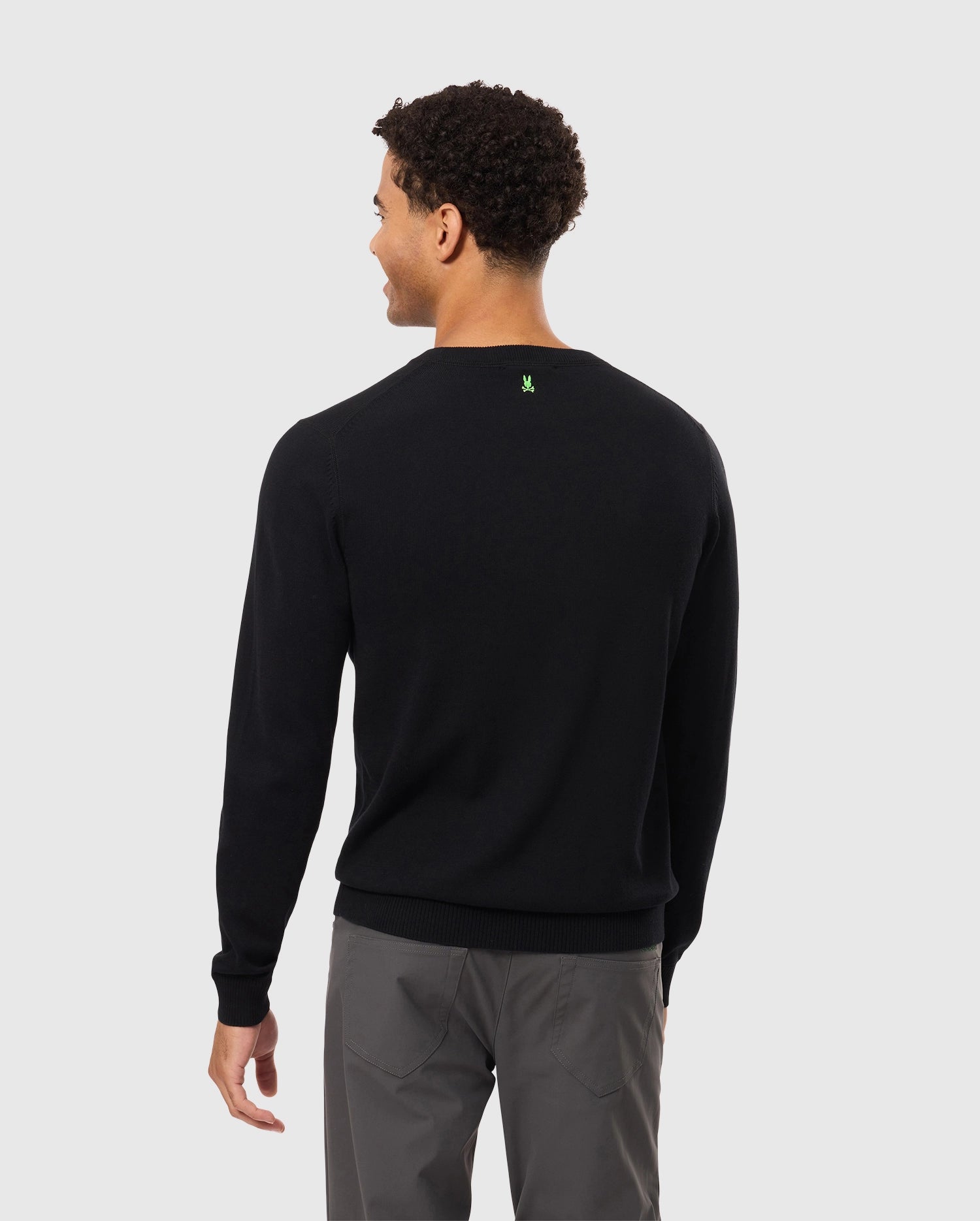 A person wearing the Psycho Bunny MENS BELMORE EXTRA FINE COTTON SWEATER in black with premium stitching and gray pants stands facing away against a plain white background.