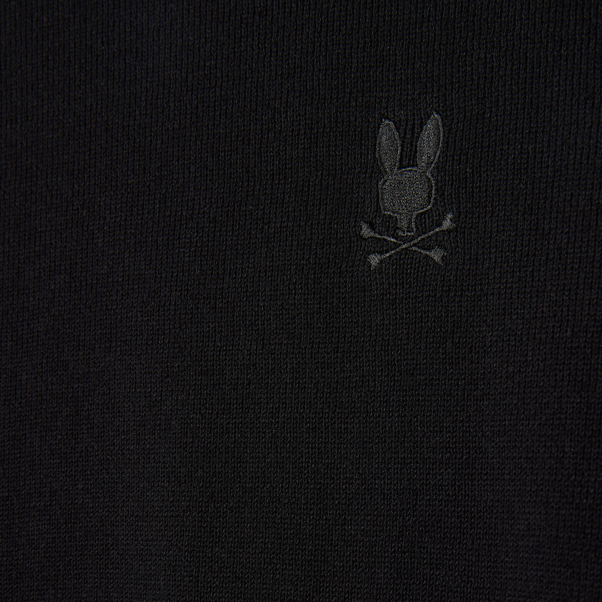 Close-up of the MENS BELMORE EXTRA FINE COTTON SWEATER by Psycho Bunny, showcasing its premium stitching and a subtly embroidered bunny logo above crossed bones on black fabric. The monochrome design seamlessly blends into the texture of extrafine cotton material.