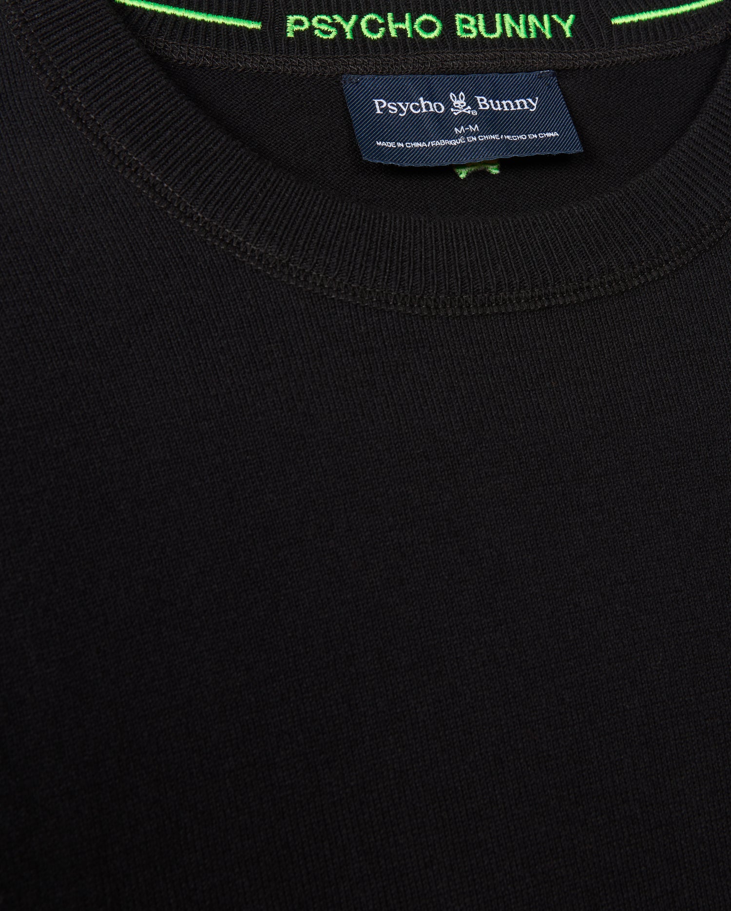 Close-up of the MENS BELMORE EXTRA FINE COTTON SWEATER - B6E832D200 by Psycho Bunny in black, highlighting its texture. The label inside displays "Psycho Bunny," with premium stitching and contrasting green thread embellishing the neckline. The tonal embroidered Bunny logo adds a subtle yet sophisticated touch.