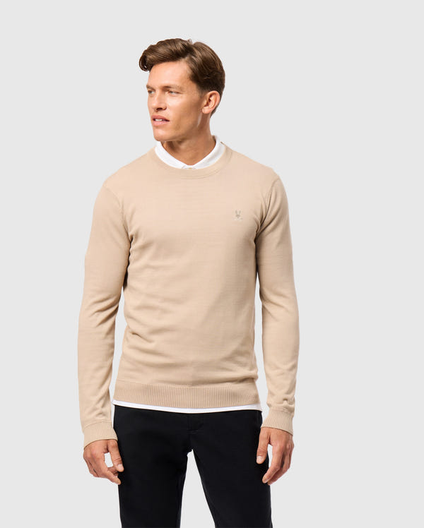 A man wears a Psycho Bunny beige MENS BELMORE EXTRA FINE COTTON SWEATER (B6E832D200) with an embroidered Bunny logo over a white collared shirt and black pants. He stands against a plain white background with short hair, looking to his right.