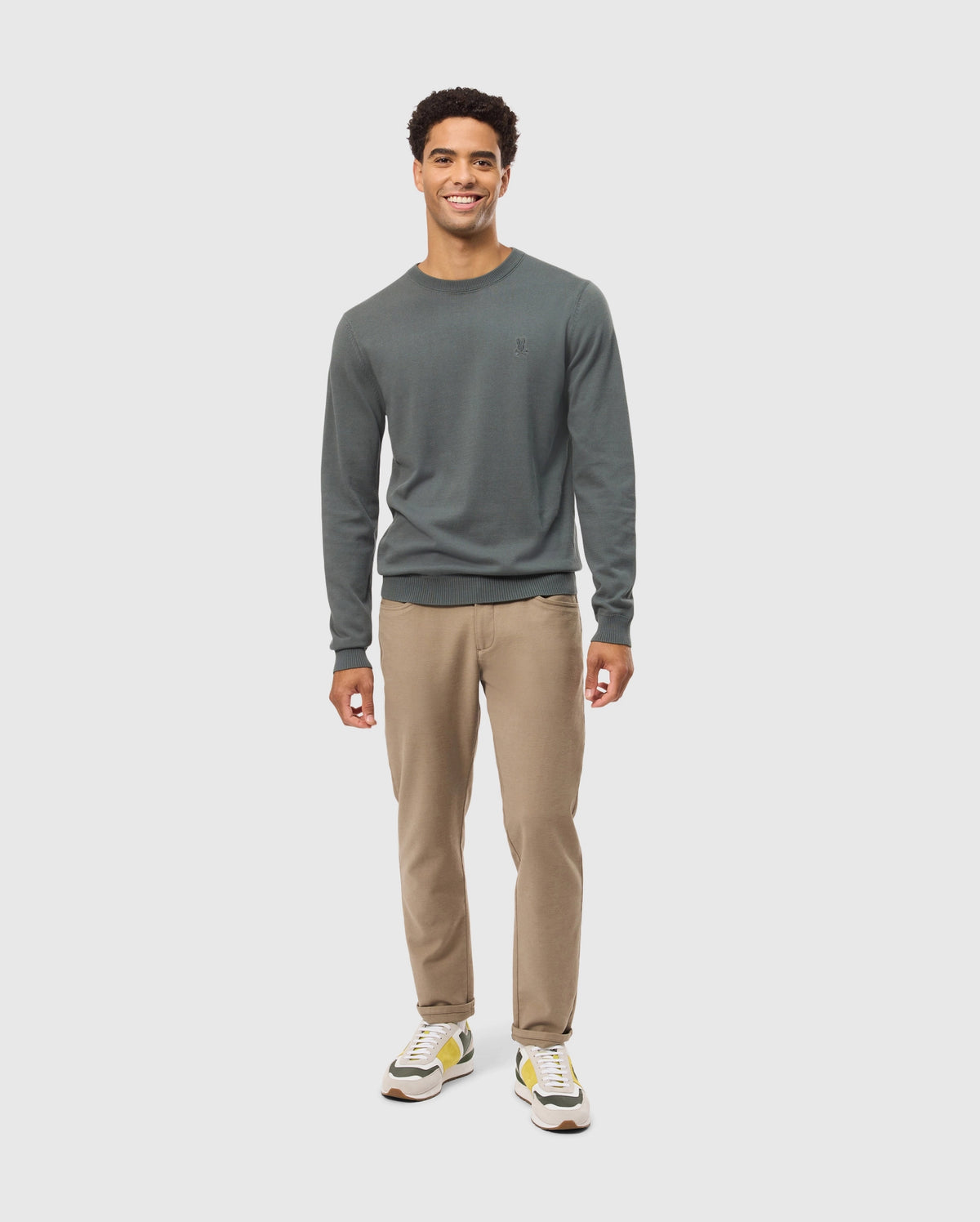 A person wearing a high-end MENS BELMORE COTTON SWEATER by Psycho Bunny, along with beige pants and white sneakers featuring yellow accents, stands against a light gray background, smiling at the camera.