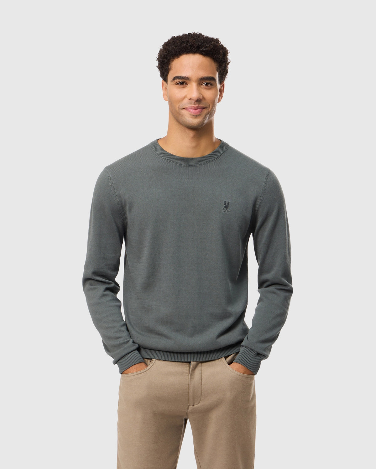 A man with short curly hair stands smiling with his hands in his pockets, wearing the high-end MENS BELMORE COTTON SWEATER by Psycho Bunny. The sweater is intricately embroidered and dark green, paired with beige pants, all set against a simple light gray background.
