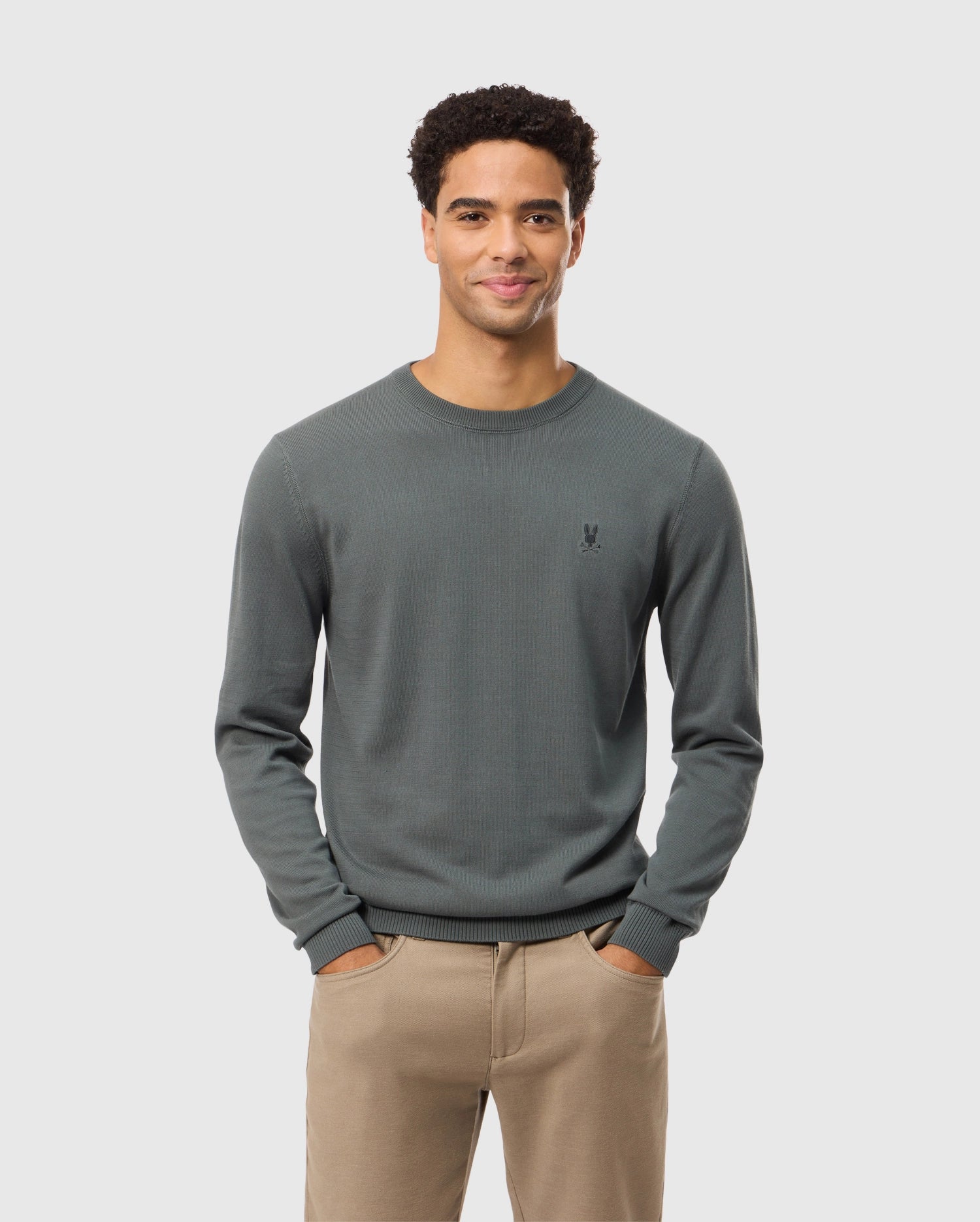 A man with short curly hair stands smiling with his hands in his pockets, wearing the high-end MENS BELMORE COTTON SWEATER by Psycho Bunny. The sweater is intricately embroidered and dark green, paired with beige pants, all set against a simple light gray background.