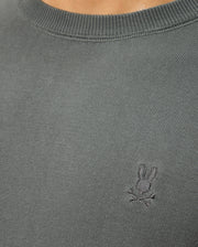Close-up of a person wearing the Psycho Bunny MENS BELMORE COTTON SWEATER - B6E832D200 in gray, featuring an embroidered design of a bunny with crossed bones on the chest.