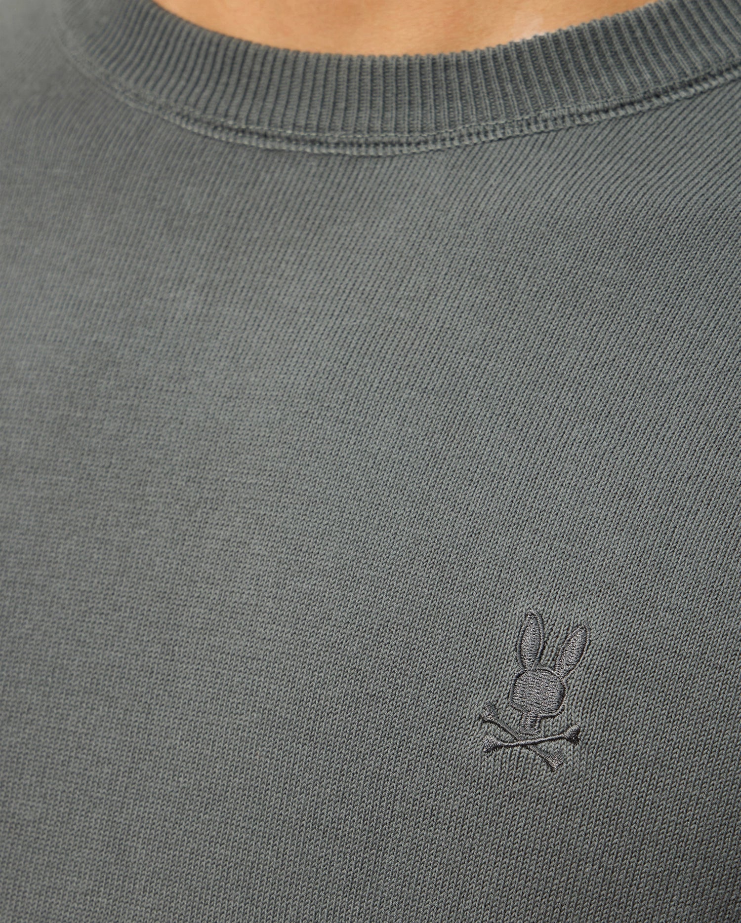 Close-up of a person wearing the Psycho Bunny MENS BELMORE COTTON SWEATER - B6E832D200 in gray, featuring an embroidered design of a bunny with crossed bones on the chest.
