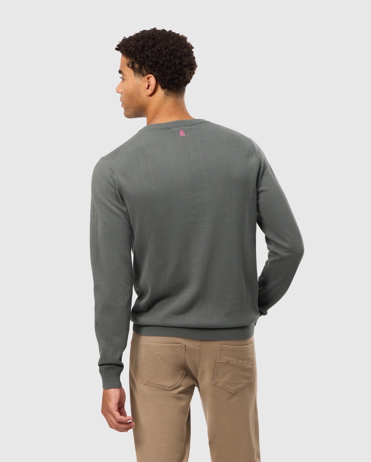A person with short curly hair, wearing the premium Psycho Bunny MEN'S BELMORE COTTON SWEATER (B6E832D200) and beige pants, stands facing away. The sweater showcases a small pink logo on the back against a plain light-colored background.