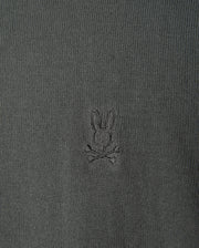 The MENS BELMORE COTTON SWEATER - B6E832D200 by Psycho Bunny features a detailed close-up of its luxurious dark green textured cotton fabric, highlighted with the brand's signature small embroidered bunny head logo above crossed bones, centered prominently on the garment.