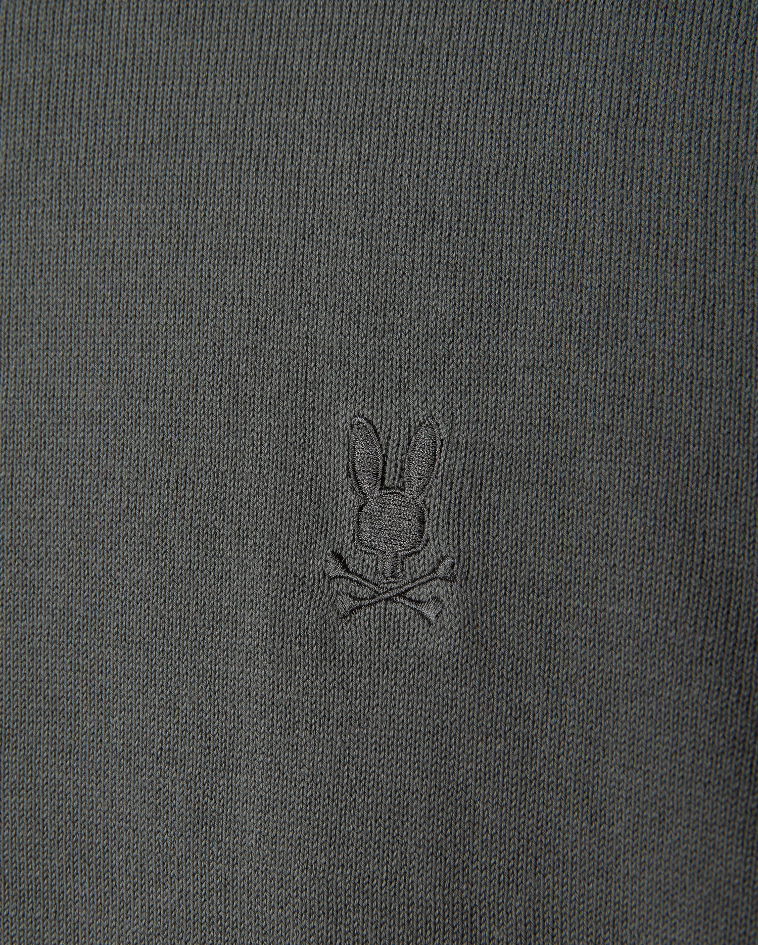 The MENS BELMORE COTTON SWEATER - B6E832D200 by Psycho Bunny features a detailed close-up of its luxurious dark green textured cotton fabric, highlighted with the brand's signature small embroidered bunny head logo above crossed bones, centered prominently on the garment.
