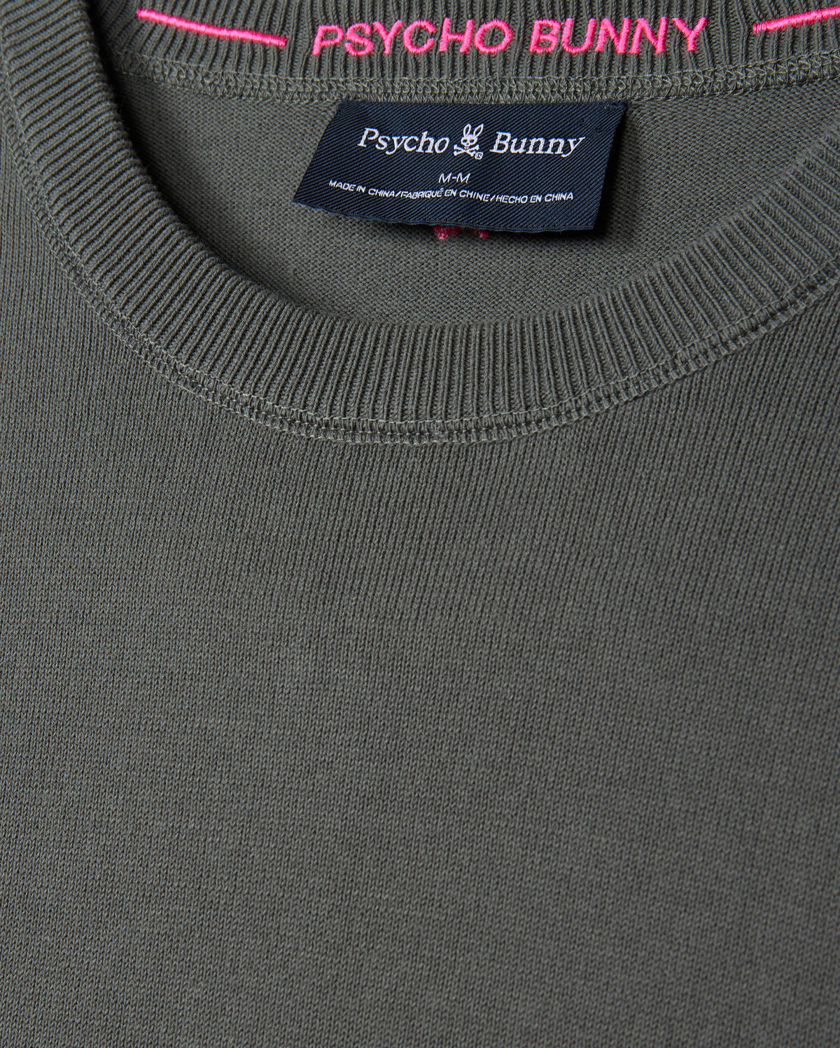 Close-up of a gray MENS BELMORE COTTON SWEATER - B6E832D200 from Psycho Bunny, showcasing a round neckline and the brand's tag inside the collar. The tag displays the Psycho Bunny logo and indicates "Made in China." The luxurious cotton pullover features pink stitching along the inside rim.