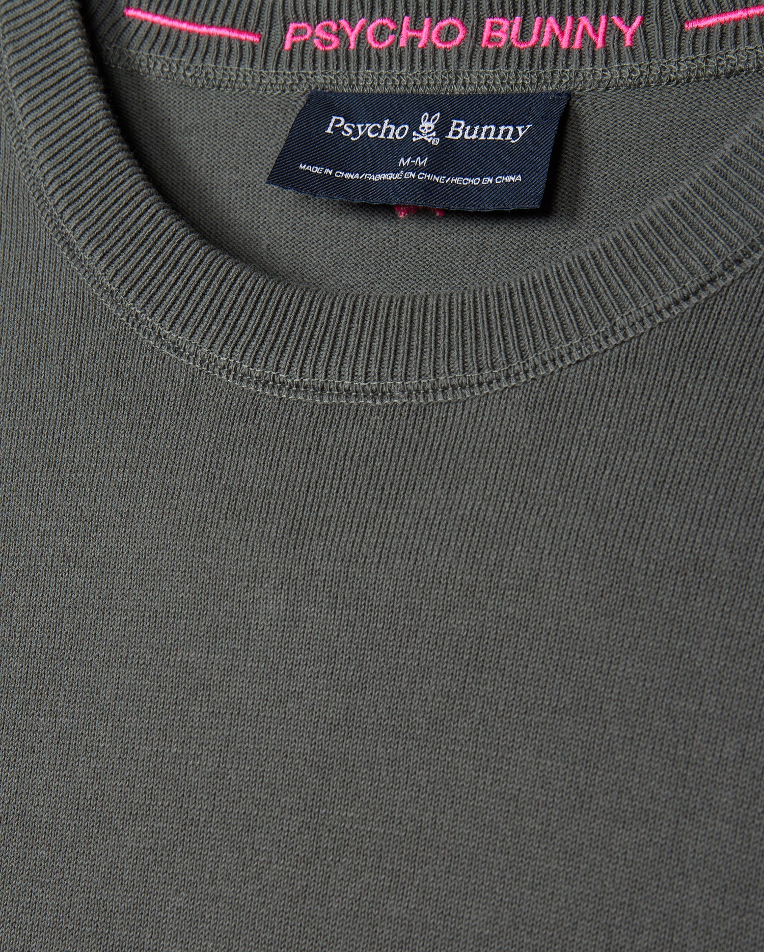 Close-up of a gray MENS BELMORE COTTON SWEATER - B6E832D200 from Psycho Bunny, showcasing a round neckline and the brand's tag inside the collar. The tag displays the Psycho Bunny logo and indicates "Made in China." The luxurious cotton pullover features pink stitching along the inside rim.