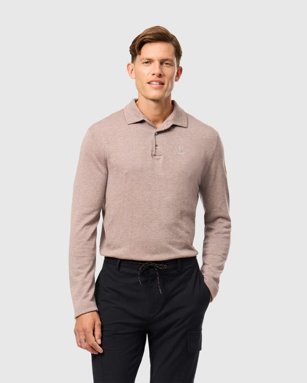 Wearing the light brown Psycho Bunny MENS BRADSHAW POLO SWEATER - B6E835D200 and black pants, a person stands against a plain gray background. They have short hair, a slight smile, and their hands at their sides.
