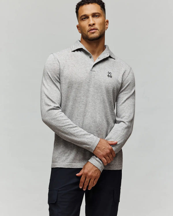 A man stands against a light gray background, confidently wearing the Psycho Bunny MENS BRADSHAW POLO SWEATER (B6E835D200) featuring a black and white bunny logo on the chest, paired with dark pants and his hands crossed in front.