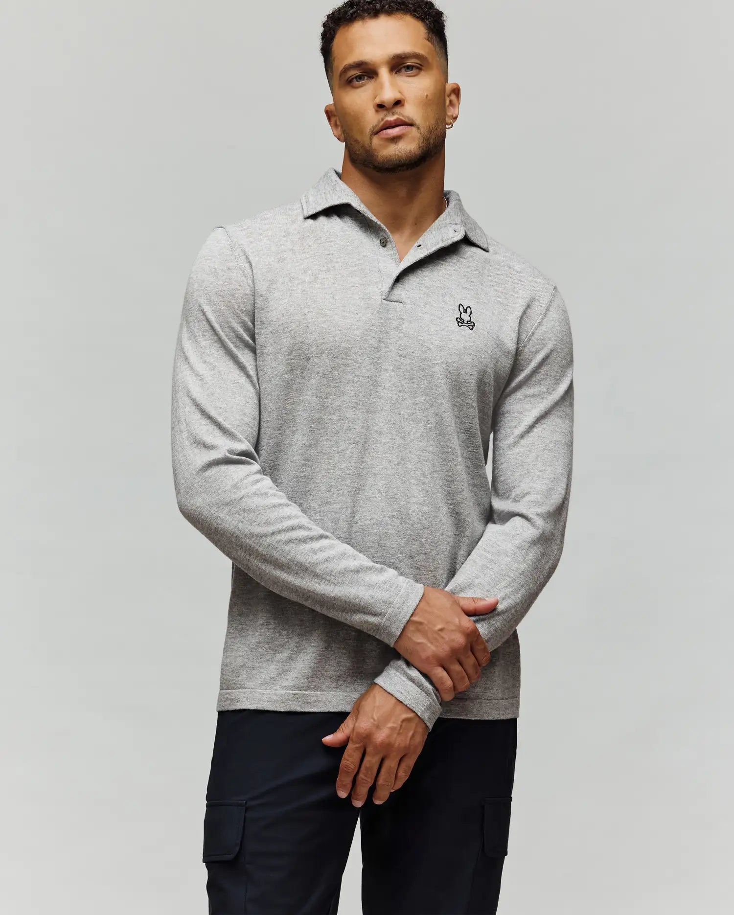 A man stands against a light gray background, confidently wearing the Psycho Bunny MENS BRADSHAW POLO SWEATER (B6E835D200) featuring a black and white bunny logo on the chest, paired with dark pants and his hands crossed in front.