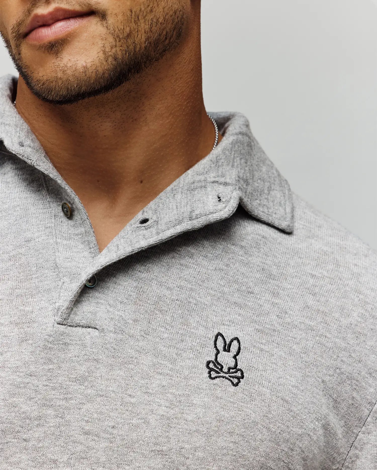 Close-up of a man in the Psycho Bunny MENS BRADSHAW POLO SWEATER (B6E835D200), light grey merino wool, featuring a small embroidered bunny head above crossbones on the chest. His face is partly visible with a trimmed beard.