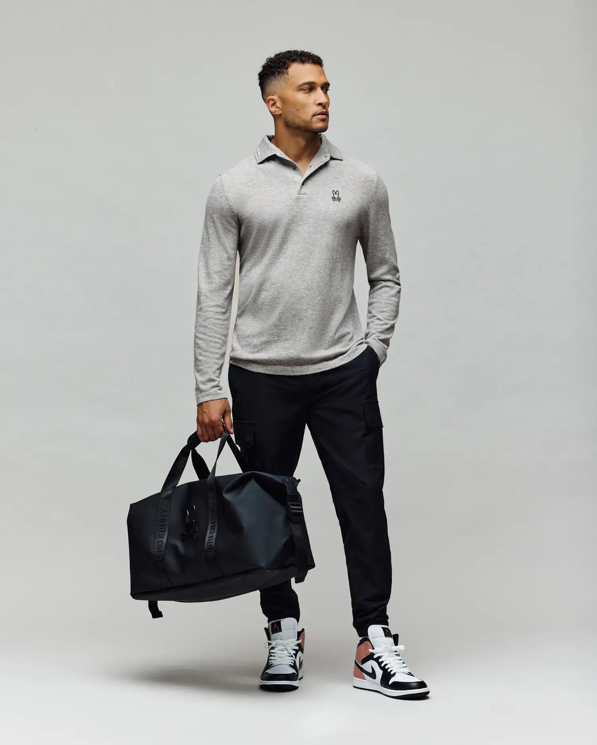 A man stands against a plain background wearing the Psycho Bunny MENS BRADSHAW POLO SWEATER in light gray with mother of pearl buttons, black pants, black-and-white sneakers, and holding a large black duffel bag in his left hand while looking right.