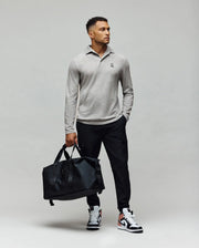 A man stands against a plain background wearing the Psycho Bunny MENS BRADSHAW POLO SWEATER in light gray with mother of pearl buttons, black pants, black-and-white sneakers, and holding a large black duffel bag in his left hand while looking right.