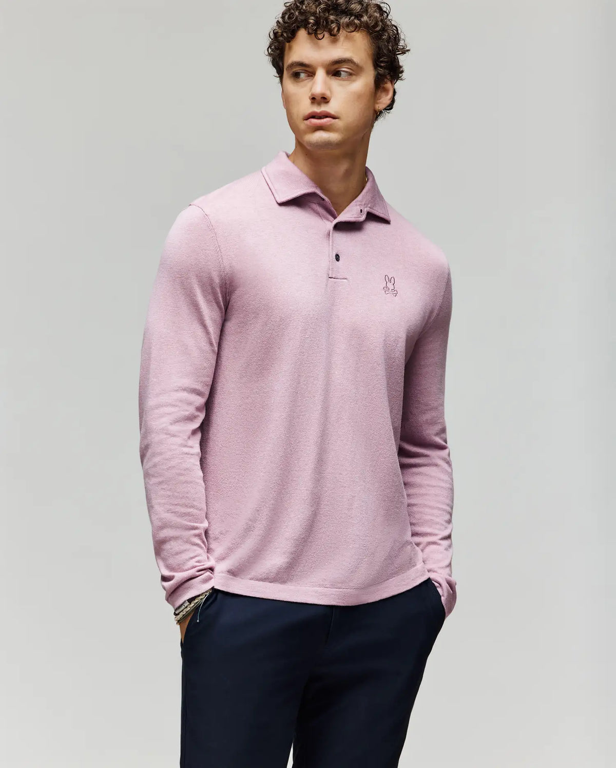 A young man with curly hair wears the Psycho Bunny MENS BRADSHAW POLO SWEATER (B6E835D200) in light pink, featuring a chest embroidered logo and mother of pearl buttons, paired with dark pants against a plain light gray background.