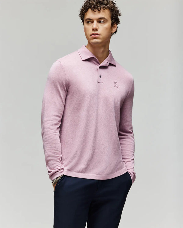 A young man with curly hair wears the Psycho Bunny MENS BRADSHAW POLO SWEATER (B6E835D200) in light pink, featuring a chest embroidered logo and mother of pearl buttons, paired with dark pants against a plain light gray background.