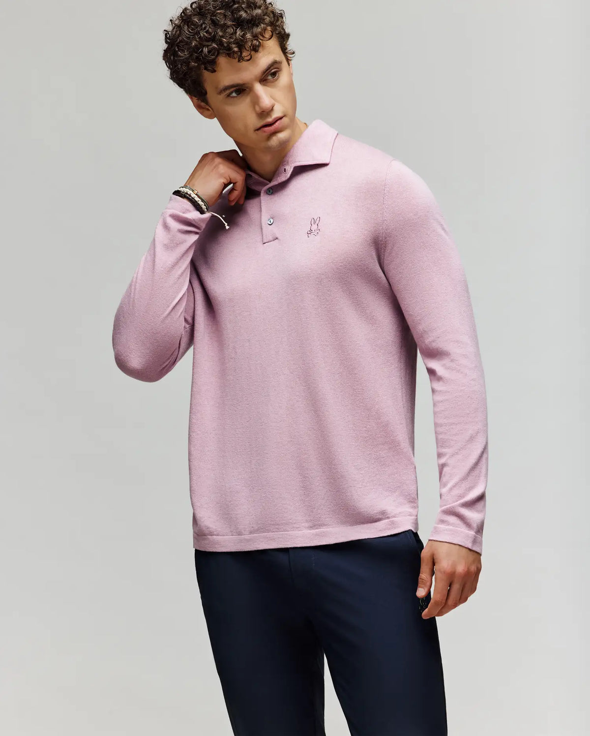 A man with curly hair wears the Psycho Bunny MENS BRADSHAW POLO SWEATER - B6E835D200 in light pink with an embroidered chest logo and dark pants, standing against a light background, touching his neck and looking to the side.