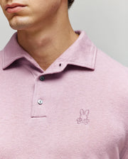 A close-up of a man wearing the Psycho Bunny MENS BRADSHAW POLO SWEATER (B6E835D200) in light purple, featuring two mother of pearl buttons and a small embroidered bunny above crossed bones logo on the chest. Only his lower face and neck are visible.