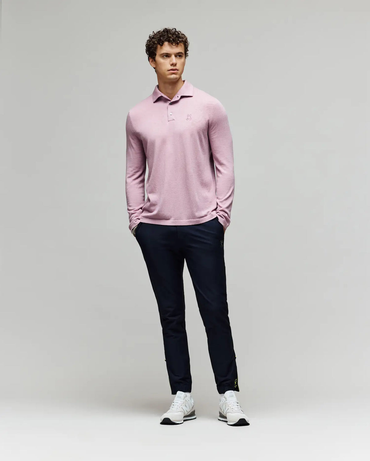 A man with curly hair stands against a plain background wearing the Psycho Bunny MENS BRADSHAW POLO SWEATER - B6E835D200 in light pink, paired with dark slim-fit pants and white sneakers, hands in pockets and looking slightly to the side.