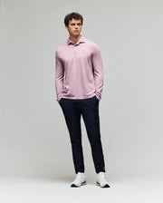 A man with curly hair stands against a plain background wearing the Psycho Bunny MENS BRADSHAW POLO SWEATER - B6E835D200 in light pink, paired with dark slim-fit pants and white sneakers, hands in pockets and looking slightly to the side.