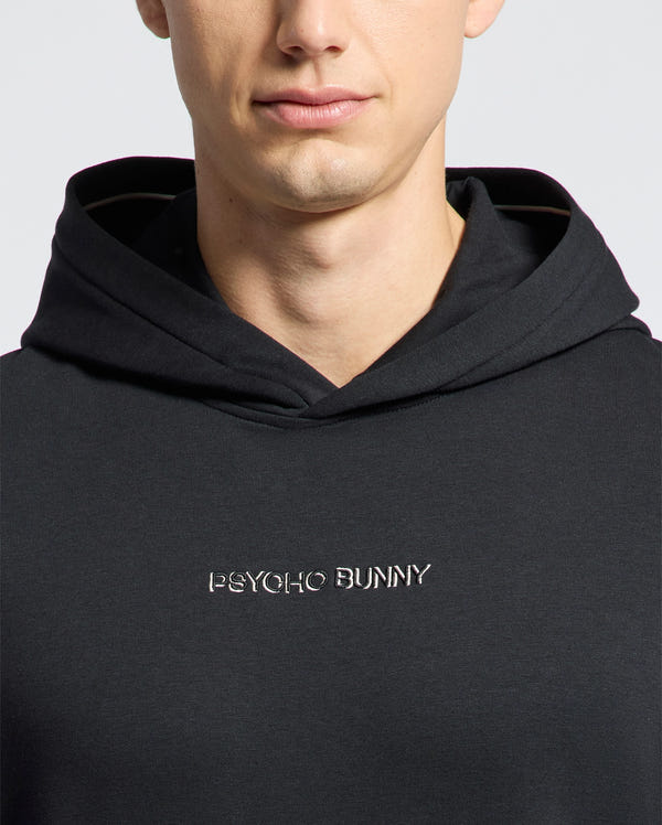 A person is wearing the MENS NORA CLASSIC FRENCH TERRY HOODIE - B6H434F200 by Psycho Bunny, a black design with "Psycho Bunny" embroidered on the front. The image shows their upper torso and partially visible face.