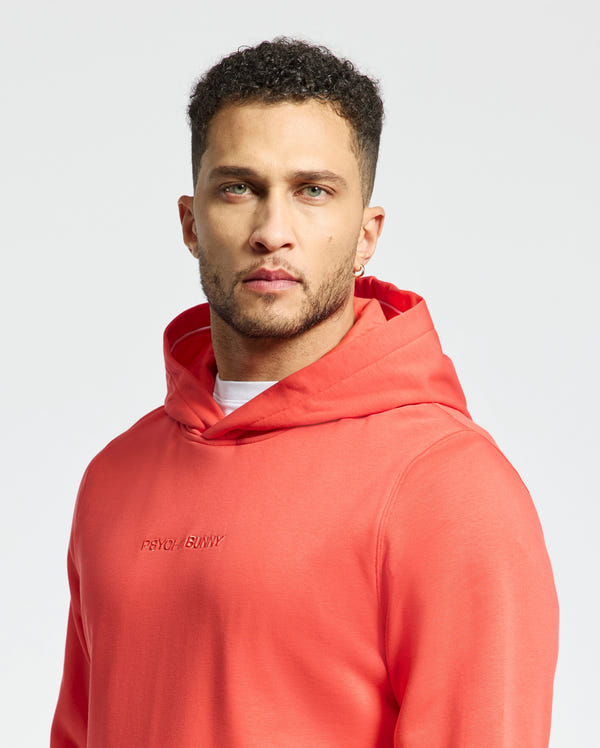 A man with short curly hair and a beard wears the coral Psycho Bunny MENS NORA CLASSIC FRENCH TERRY HOODIE - B6H434F200, showcasing an embroidered wordmark, as he stands against a plain white background.