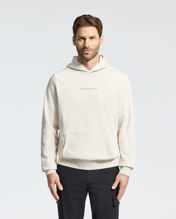 A bearded man with short hair stands against a plain background, wearing a light-colored MENS NORA CLASSIC FRENCH TERRY HOODIE by Psycho Bunny. "PSYCHO BUNNY" and its embroidered logo are displayed on the hoodie. He pairs it with dark pants, his hands calmly at his sides, expression neutral.