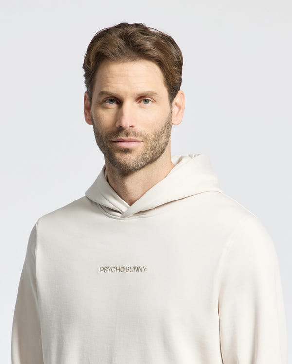 A man with short brown hair and a beard wears the light-colored Psycho Bunny MENS NORA CLASSIC FRENCH TERRY HOODIE - B6H434F200, featuring an embroidered logo. The plain gray background accentuates its stylish, casual vibe crafted from soft French terry.