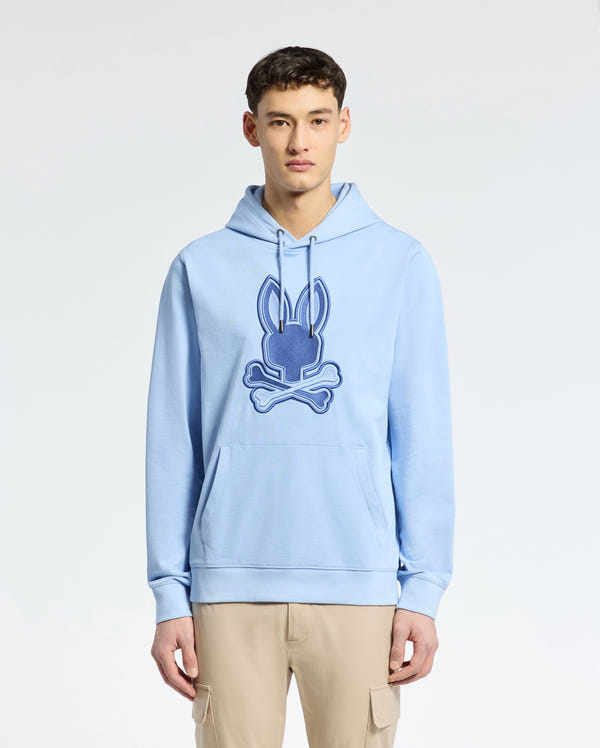 A person in a light blue Psycho Bunny MENS NEW CASTLE EMBROIDERED FRENCH TERRY HOODIE, showcasing a Bunny skull logo, stands against a plain background. They're paired with beige cargo pants, experiencing the soft comfort of French terry fabric.