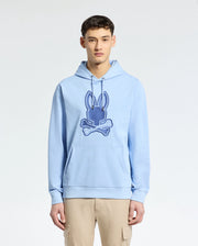A person in a light blue Psycho Bunny MENS NEW CASTLE EMBROIDERED FRENCH TERRY HOODIE, showcasing a Bunny skull logo, stands against a plain background. They're paired with beige cargo pants, experiencing the soft comfort of French terry fabric.
