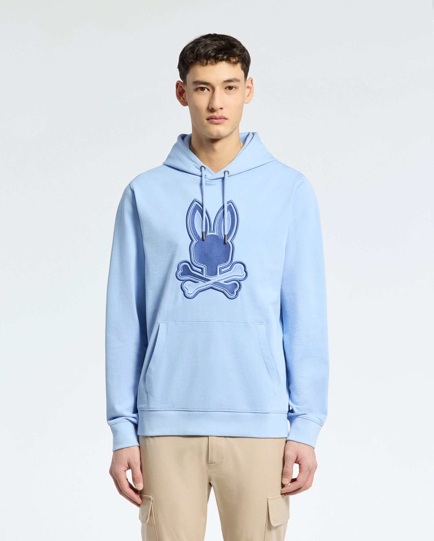 A person in a light blue Psycho Bunny MENS NEW CASTLE EMBROIDERED FRENCH TERRY HOODIE, showcasing a Bunny skull logo, stands against a plain background. They're paired with beige cargo pants, experiencing the soft comfort of French terry fabric.