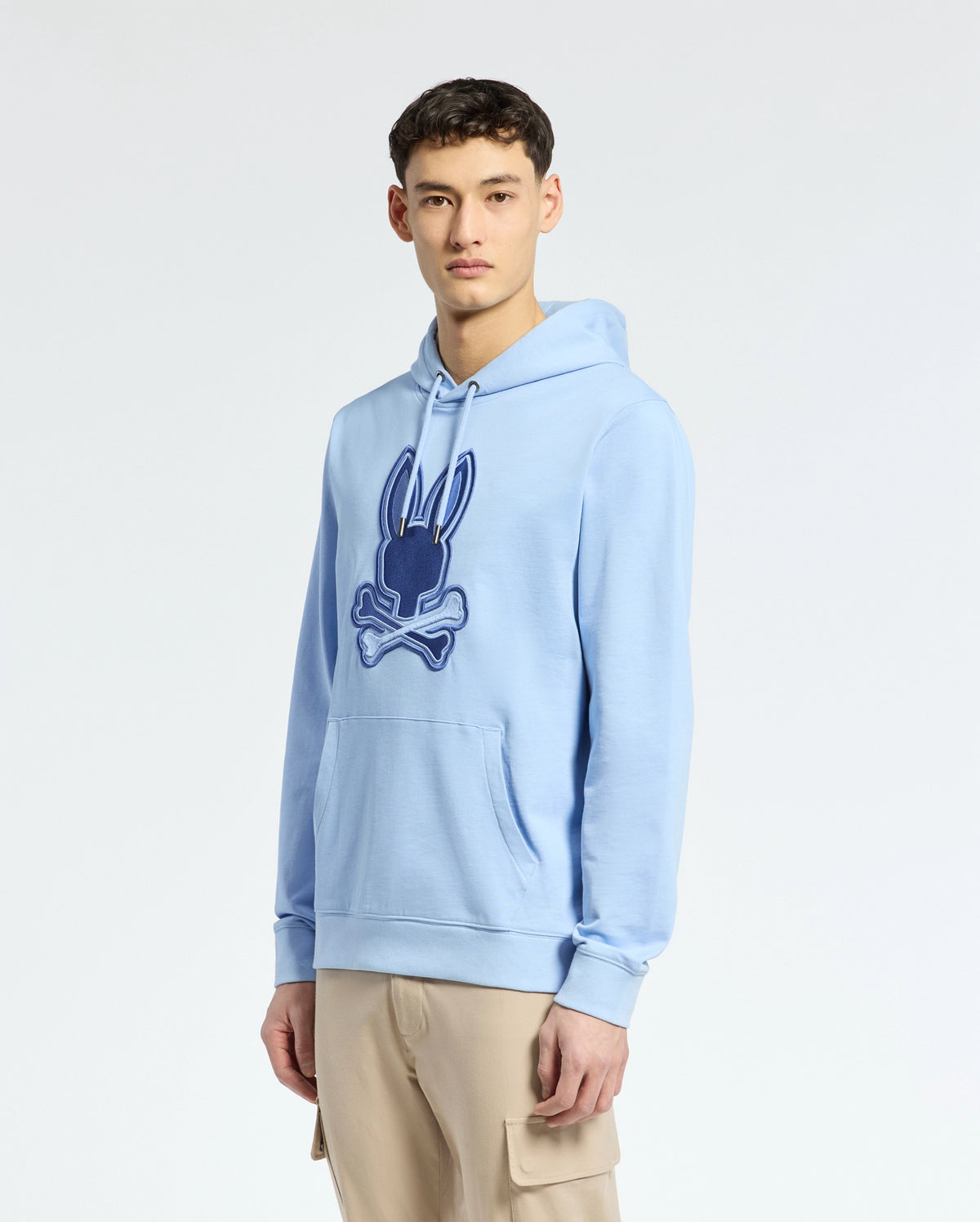 A young man wears a Psycho Bunny light blue MENS NEW CASTLE EMBROIDERED FRENCH TERRY HOODIE, showcasing the embroidered bunny emblem on the chest. He pairs it with beige cargo pants against a plain white background.