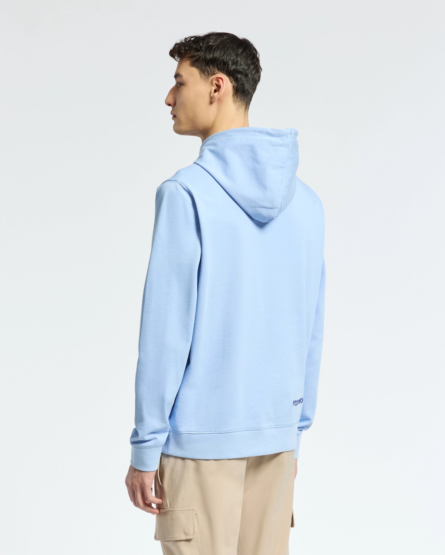 A person stands slightly left, wearing a Psycho Bunny's Mens New Castle Embroidered French Terry Hoodie (B6H517F200) in light blue with beige pants. The hood is down against a plain white backdrop, showing the side and back view.