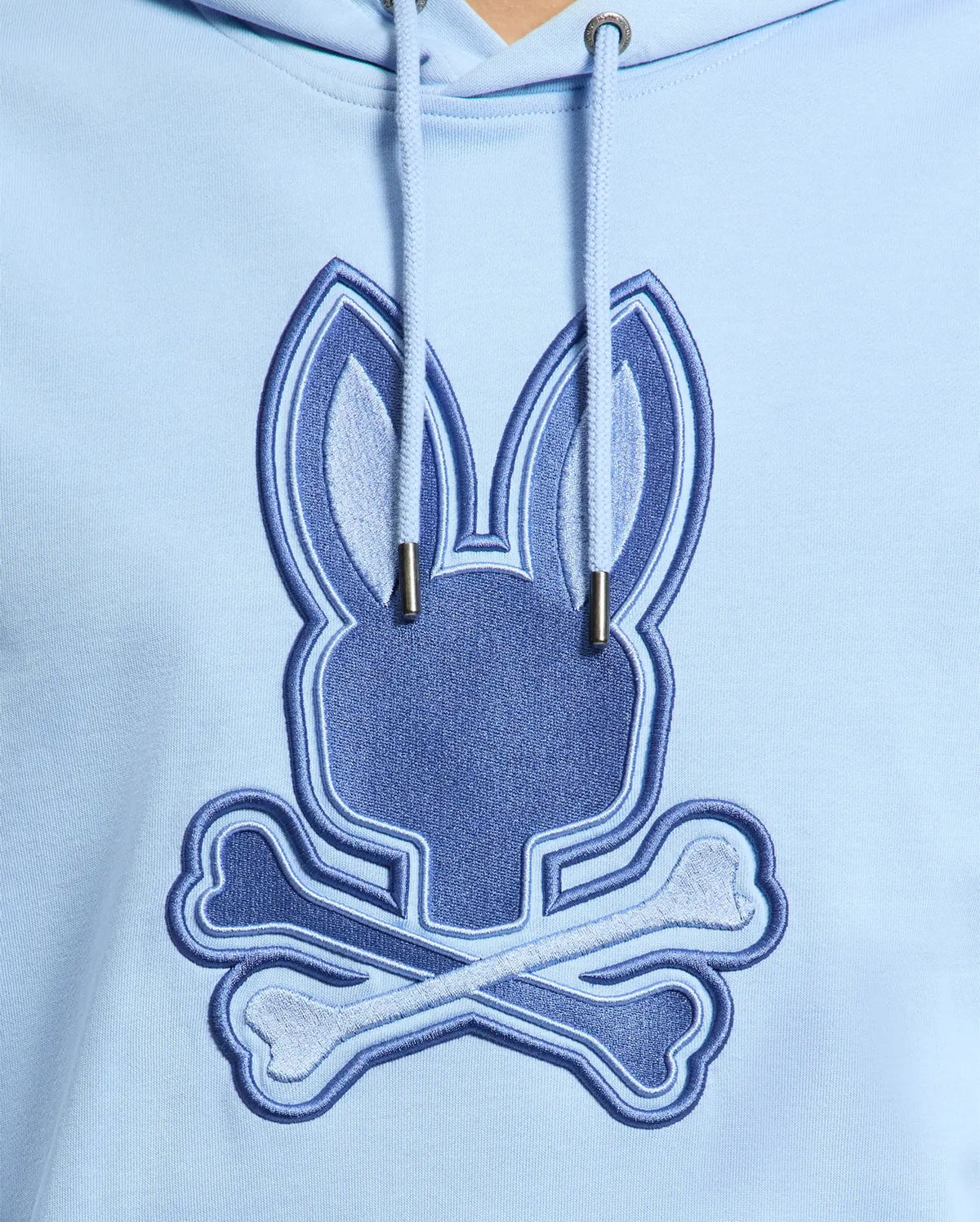 The Psycho Bunny Men's New Castle Hoodie in light blue features an embroidered bunny head with tall ears and crossed bones, resembling a playful skull and crossbones. Made from cozy French terry fabric, it includes prominent strings for added style. Model: B6H517F200.