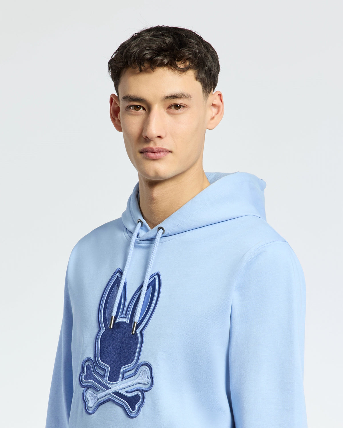 A person wears a light blue Psycho Bunny New Castle hoodie (B6H517F200), featuring a dark blue embroidered bunny head with crossed bones. The French terry fabric adds casual elegance against a plain, light background.