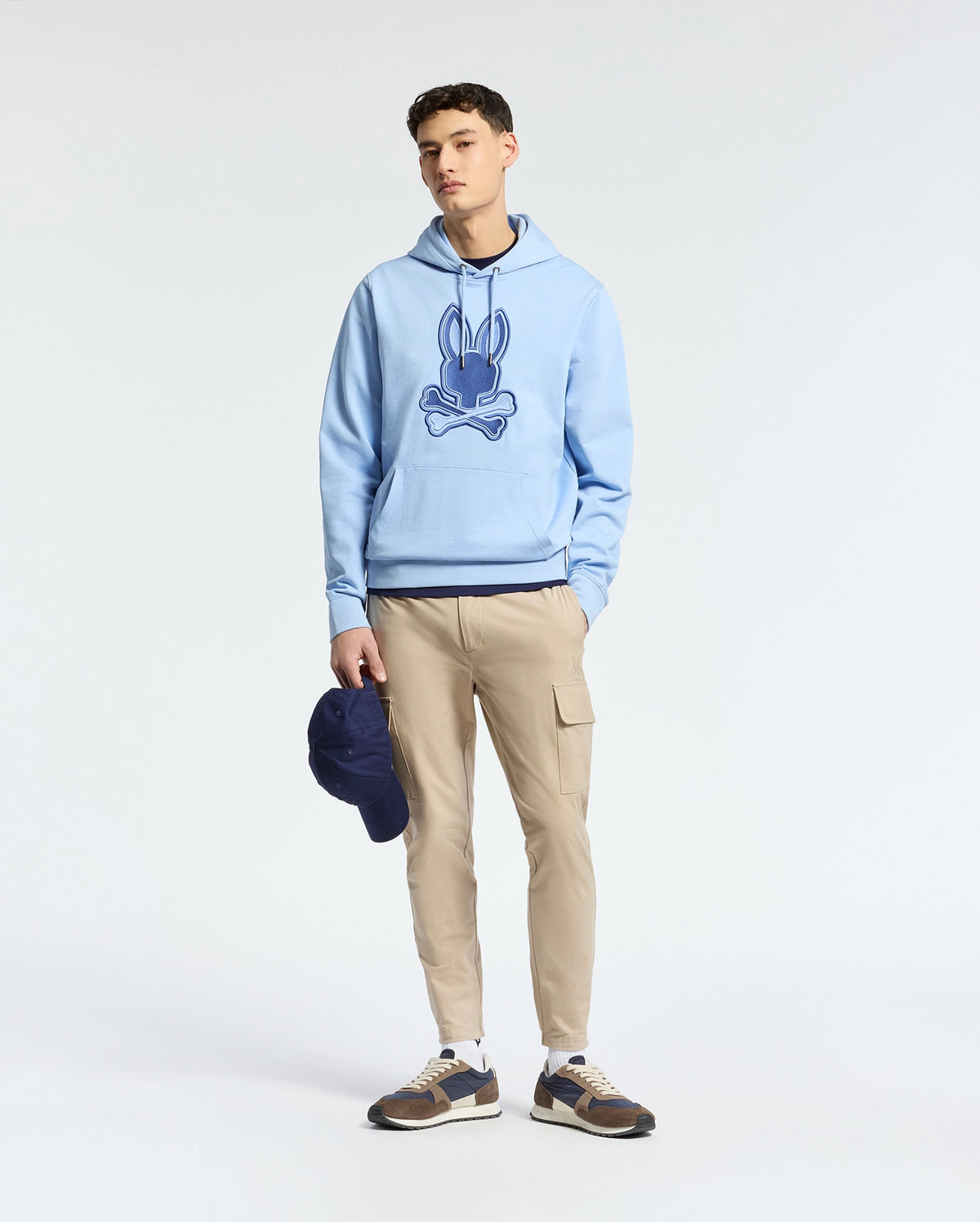 A person in a light blue Psycho Bunny MENS NEW CASTLE EMBROIDERED FRENCH TERRY HOODIE - B6H517F200 stands against a neutral background, wearing beige cargo pants and brown sneakers, holding a dark blue cap in their left hand.