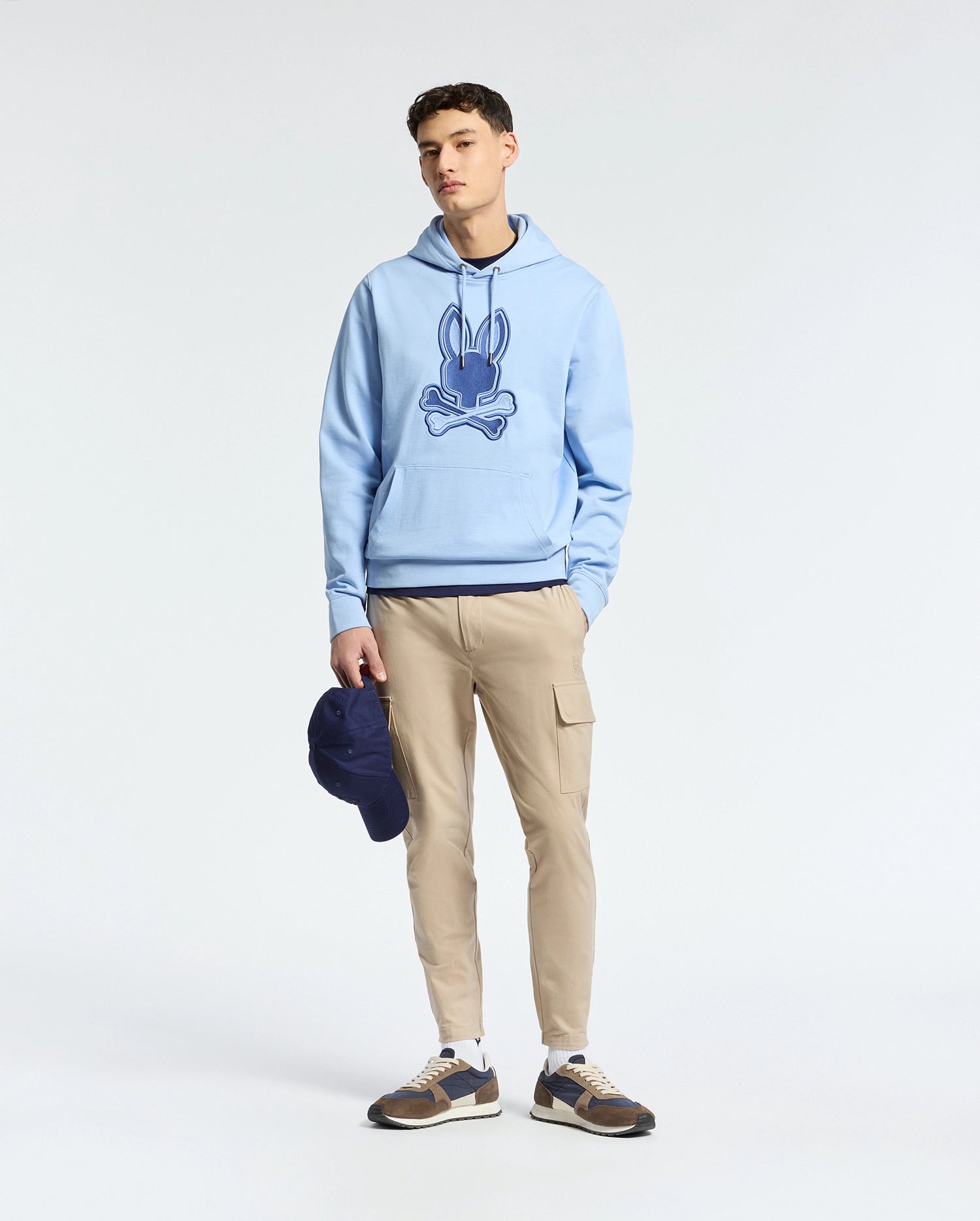 A person in a light blue Psycho Bunny MENS NEW CASTLE EMBROIDERED FRENCH TERRY HOODIE - B6H517F200 stands against a neutral background, wearing beige cargo pants and brown sneakers, holding a dark blue cap in their left hand.