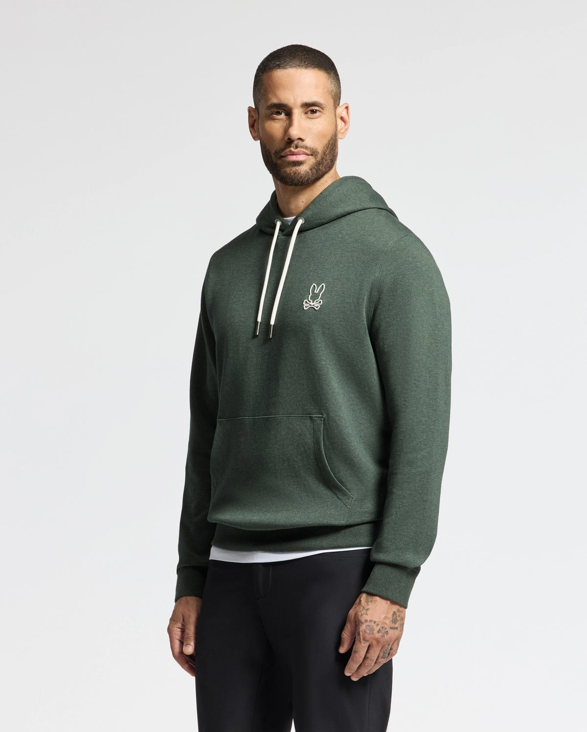 A man wearing the stylish MENS JAMES PREMIUM HOODIE - B6H789D200 by Psycho Bunny, featuring a petite rabbit logo on the chest, stands against a plain background. This high-quality men's cotton hoodie boasts white drawstrings and is paired with black pants.