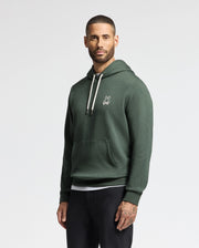 A man wearing the stylish MENS JAMES PREMIUM HOODIE - B6H789D200 by Psycho Bunny, featuring a petite rabbit logo on the chest, stands against a plain background. This high-quality men's cotton hoodie boasts white drawstrings and is paired with black pants.