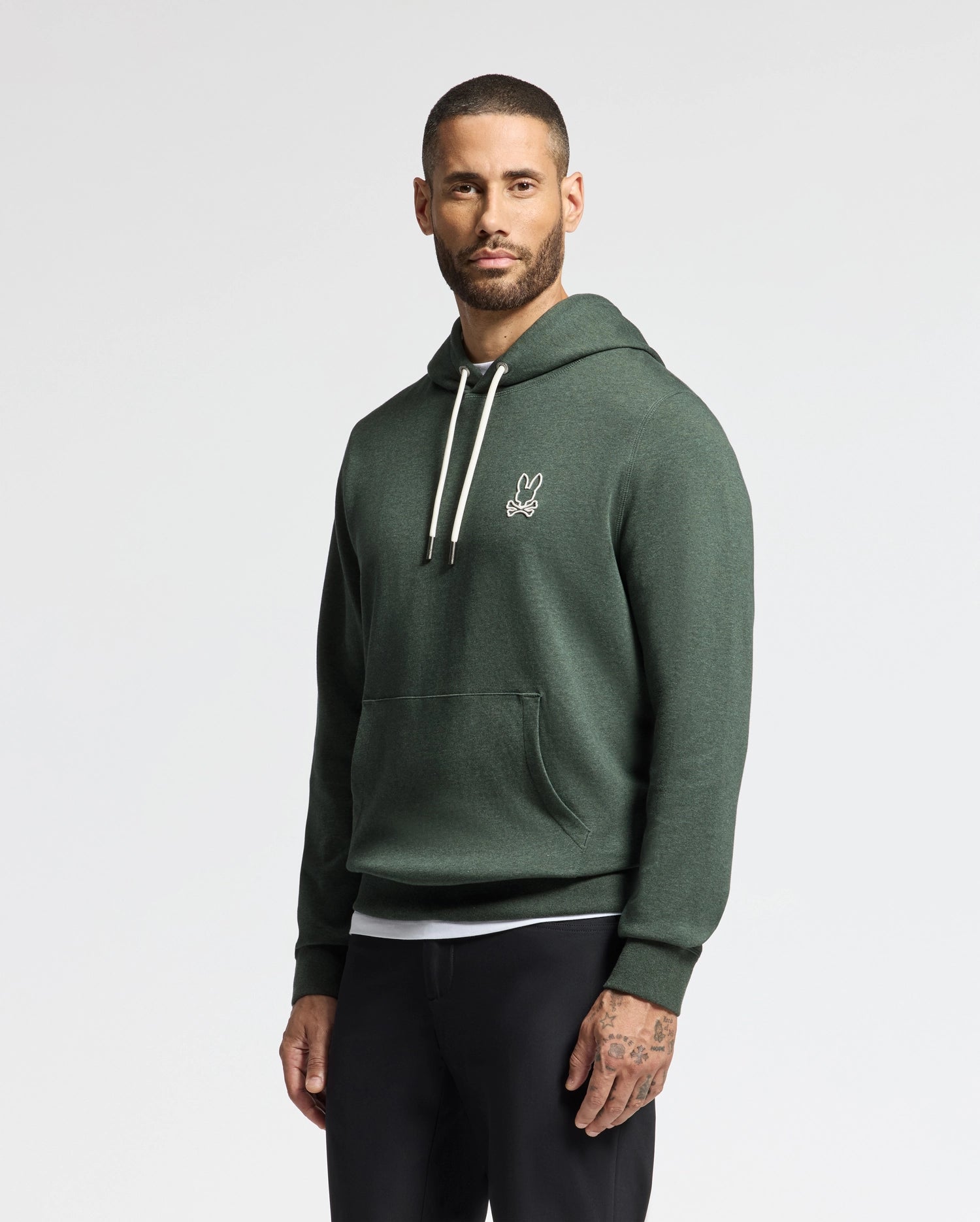 A man wearing the stylish MENS JAMES PREMIUM HOODIE - B6H789D200 by Psycho Bunny, featuring a petite rabbit logo on the chest, stands against a plain background. This high-quality men's cotton hoodie boasts white drawstrings and is paired with black pants.