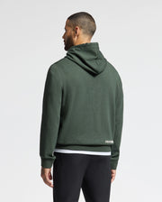 A person stands with their back to the camera, wearing the Psycho Bunny Men's James Premium Hoodie (B6H789D200) in dark green, with the hood down and paired with black pants. The plain white background accentuates the stylish simplicity of this lightweight hoodie for men.