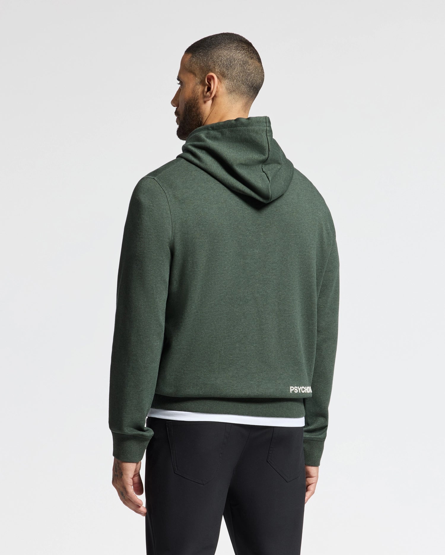 A person stands with their back to the camera, wearing the Psycho Bunny Men's James Premium Hoodie (B6H789D200) in dark green, with the hood down and paired with black pants. The plain white background accentuates the stylish simplicity of this lightweight hoodie for men.