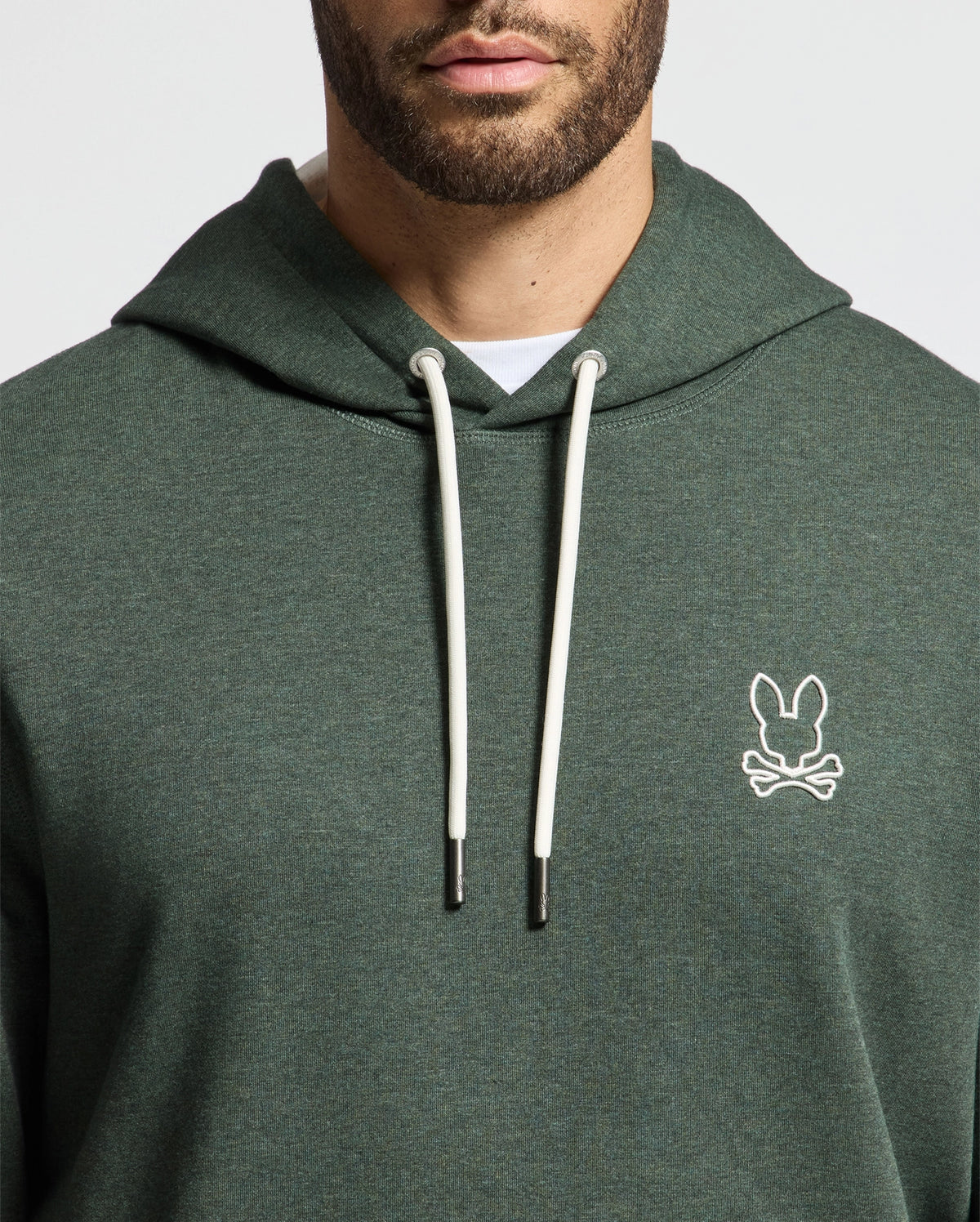 A man wears the MENS JAMES PREMIUM HOODIE - B6H789D200 by Psycho Bunny, showcasing a stylish green design with a distinctive white rabbit and crossbones logo on the chest. This high-quality men's cotton hoodie features white drawstrings and is paired with a white t-shirt underneath for a complete look.