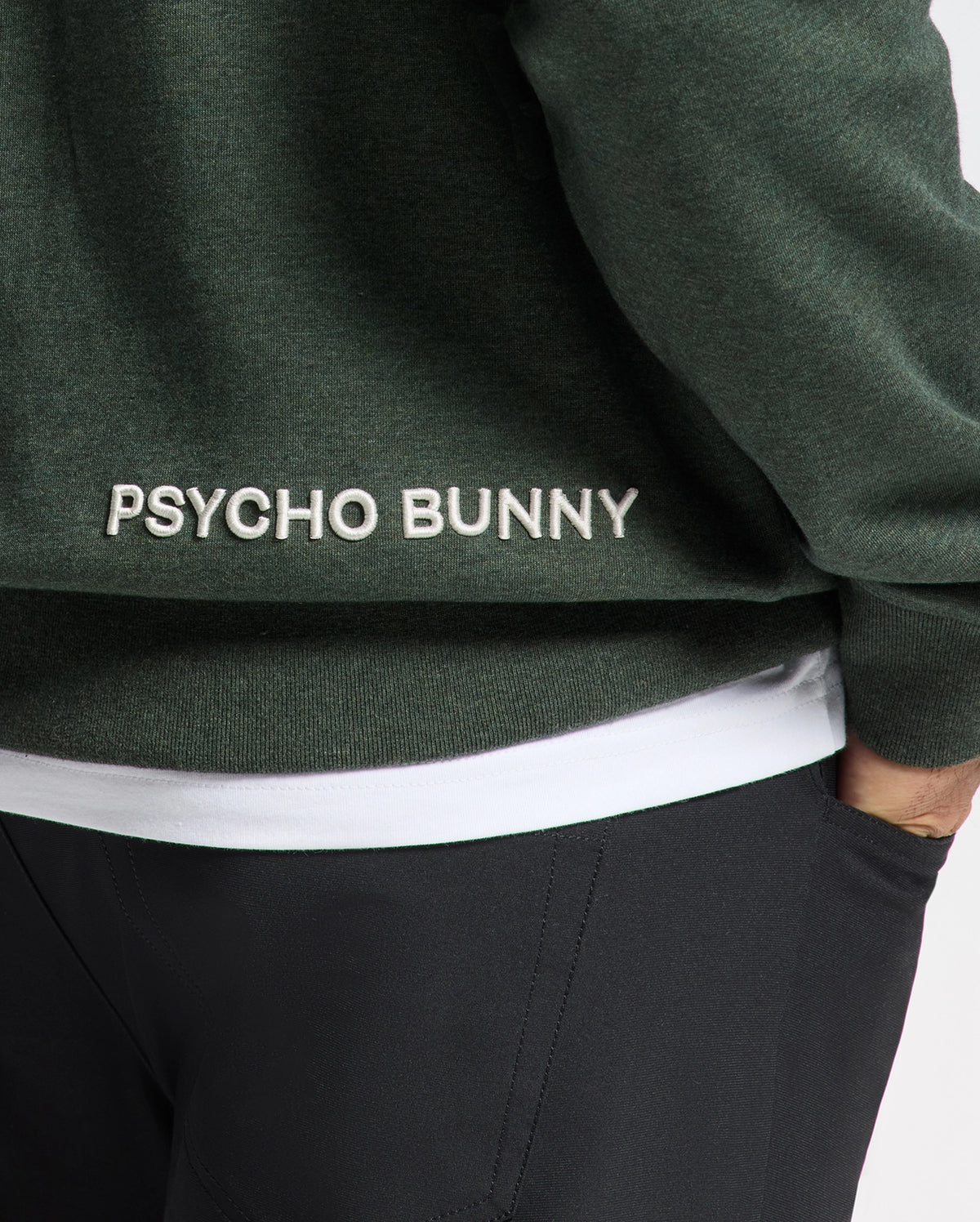 A close-up of a person wearing the MENS JAMES PREMIUM HOODIE - B6H789D200, a lightweight green hoodie designed for men by Psycho Bunny, featuring "Psycho Bunny" embroidered in white on the back. Paired with black pants and a white shirt underneath, this stylish hooded sweatshirt exudes effortless style.
