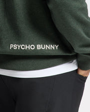 A close-up of a person wearing the MENS JAMES PREMIUM HOODIE - B6H789D200, a lightweight green hoodie designed for men by Psycho Bunny, featuring "Psycho Bunny" embroidered in white on the back. Paired with black pants and a white shirt underneath, this stylish hooded sweatshirt exudes effortless style.