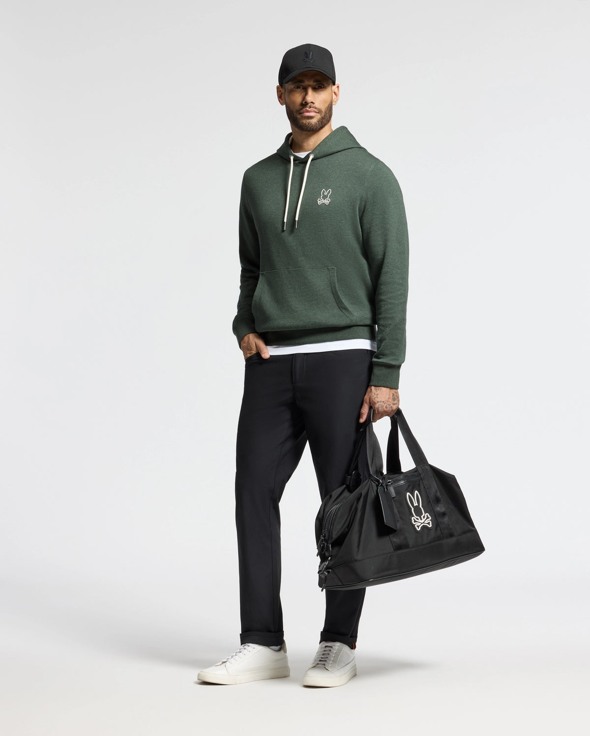 A man stands against a plain backdrop wearing the trendy MENS JAMES PREMIUM HOODIE in green by Psycho Bunny, paired with black pants and white sneakers. He holds a black duffel bag featuring the Psycho Bunny logo in his left hand and wears a black cap.