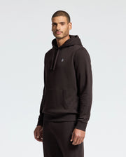 A man showcases the Psycho Bunny MENS LEVY ESSENTIAL FLEECE HOODIE - B6H859D200, made from soft French terry. This black hoodie, composed of 100% cotton, is adorned with a small embroidered logo on the chest. He poses against a plain white backdrop, facing forward with a neutral expression. The hoodie features a front pocket and drawstrings.